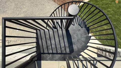 Overhead view of a black spiral staircase with metal railings casting shadows on concrete and grass