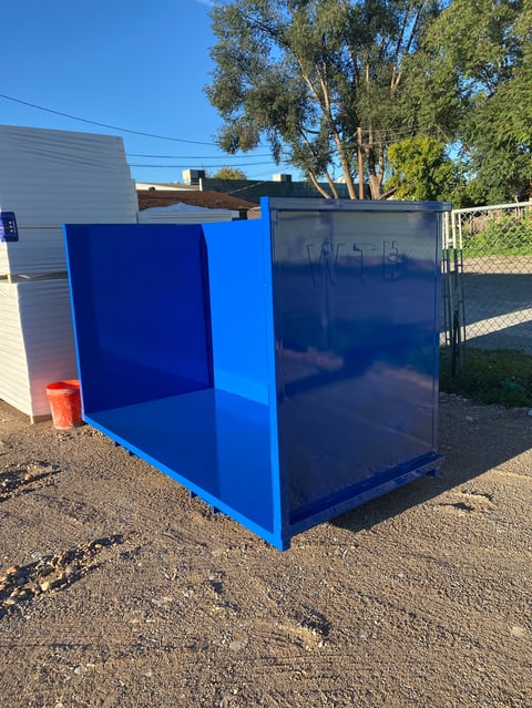 Bright blue metal storage box or shipping container positioned in a dirt lot with trees and buildings in the background