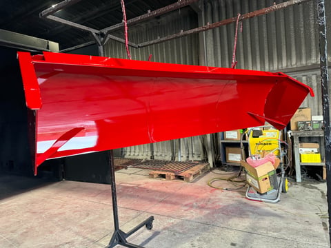 Large red metal panel or boat hull hanging from ceiling in industrial workshop with concrete floor and corrugated metal walls