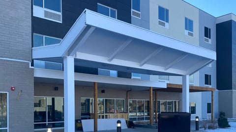 Modern building entrance with white metal canopy, glass doors and windows, and gray stone and dark paneled facade