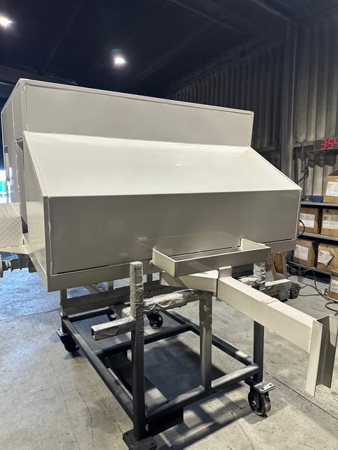 Large stainless steel industrial equipment with angled white hood mounted on black wheeled cart in warehouse