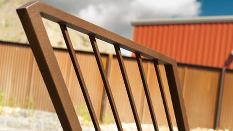 Close-up of a brown metal fence panel with vertical bars against a blurred corrugated metal building background